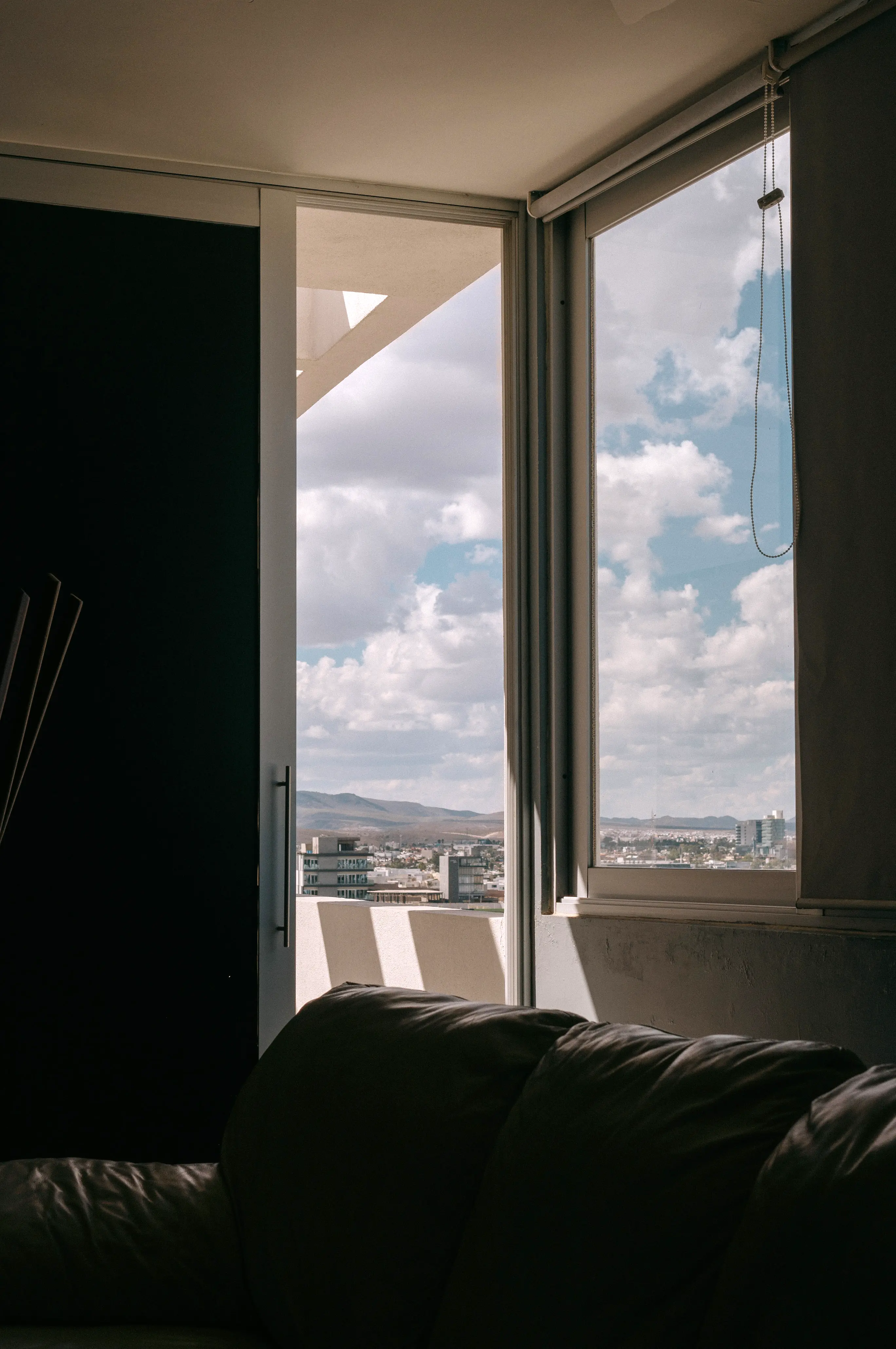 Foto de una habitación. A través de la ventana se observa un gran cielo nuboso a la distancia. La habitación está a oscuras, iluminada por la luz remanante de la ventana