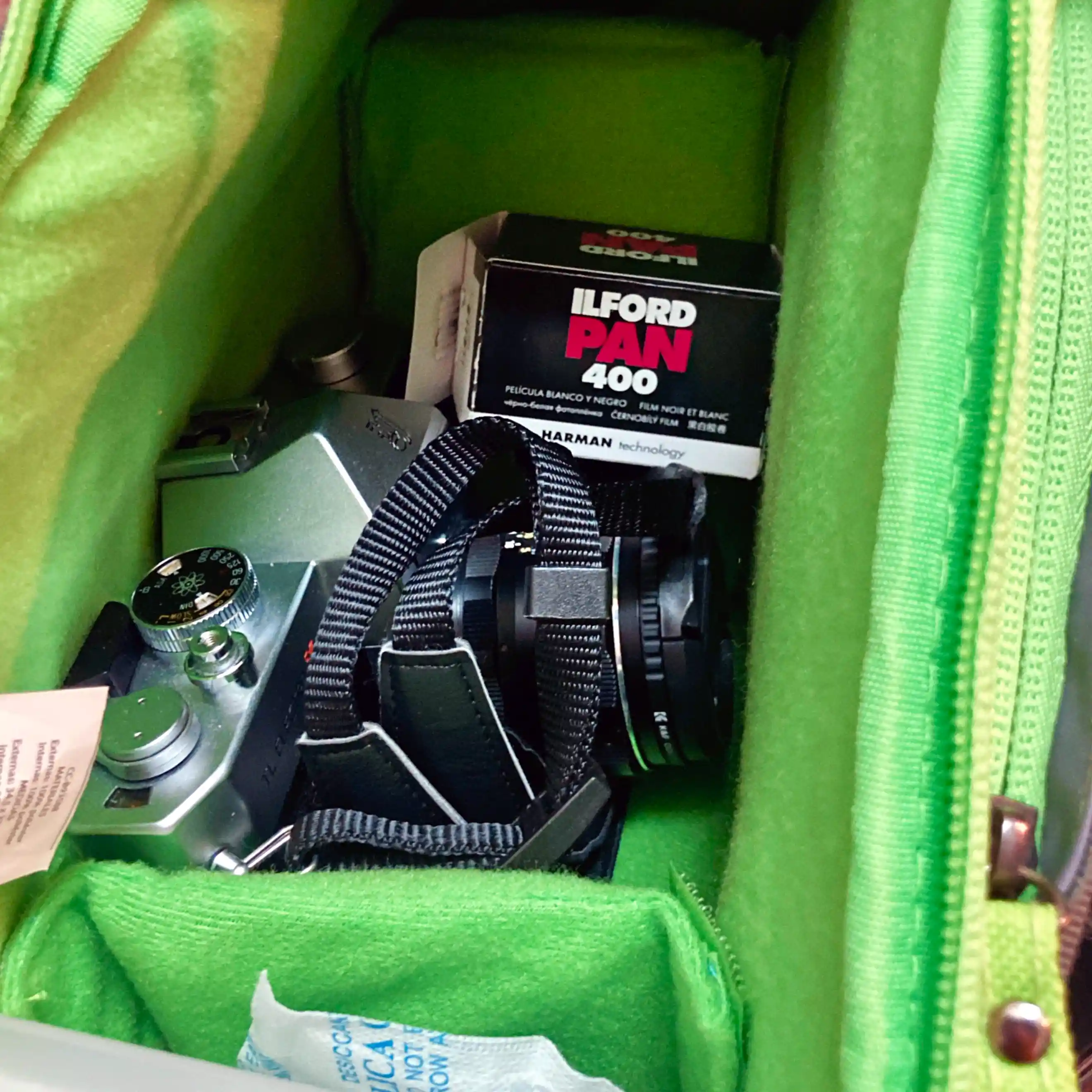 Una cámara analógica con su correa envuelta sobre ella dentro de una mochila fotográfica. Al fondo se observa una caja de película Ilford